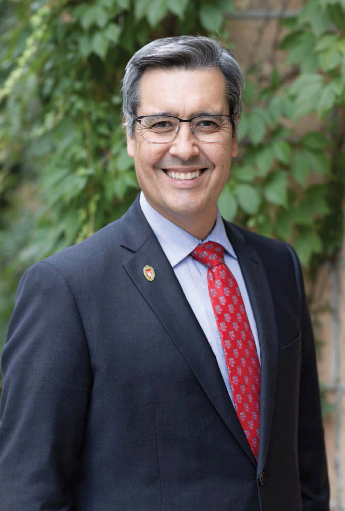 UW Law School Dean, Dan Tokaji, stands in front of a brick wall covered in leafy vines. He smiles to the camera.