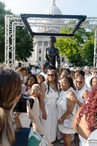 A smiling group of people, including a lot of people of color, standing in front of the Vel Phillips statue, smiling, and all wearing white.