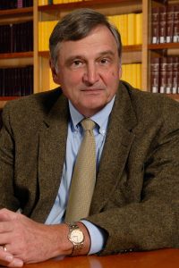 Headshot photo of David Schultz looking into the camera. He is wearing a brown wool suit jacket, blue shirt, and beige tie. His silver and gold watch shines on his wrist.