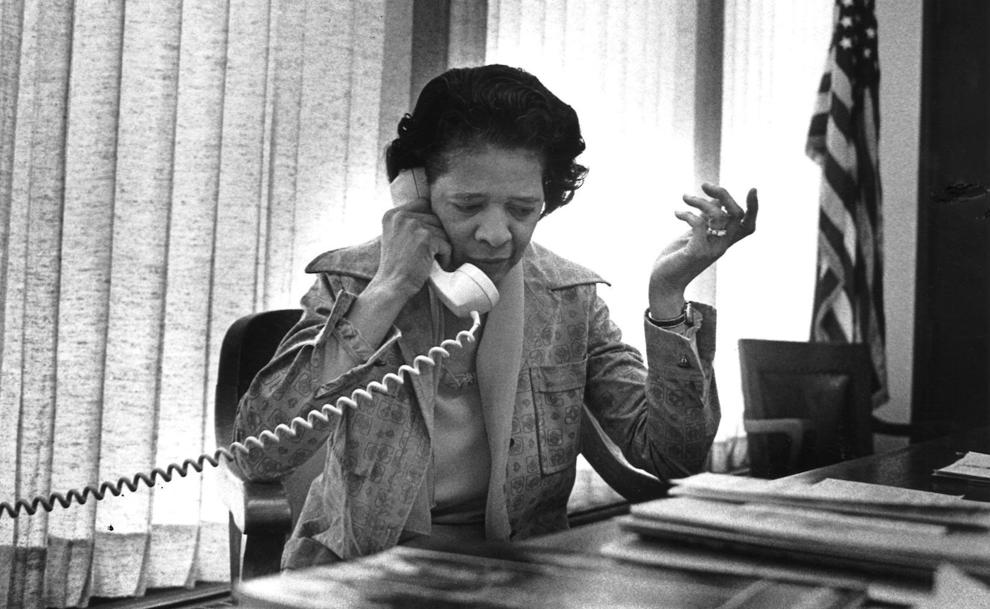 Black and white photo of Vel Phillips using a corded telephone while seated at a desk in front of a shaded window and an American flag on a pole in the corner.