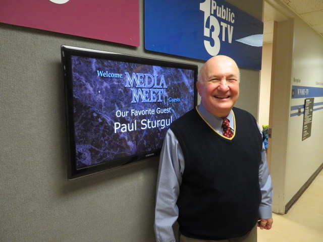 Photo of Paul Sturgul smiling as he stands in front of a screen displaying his name as a guest on a Media Meet event.