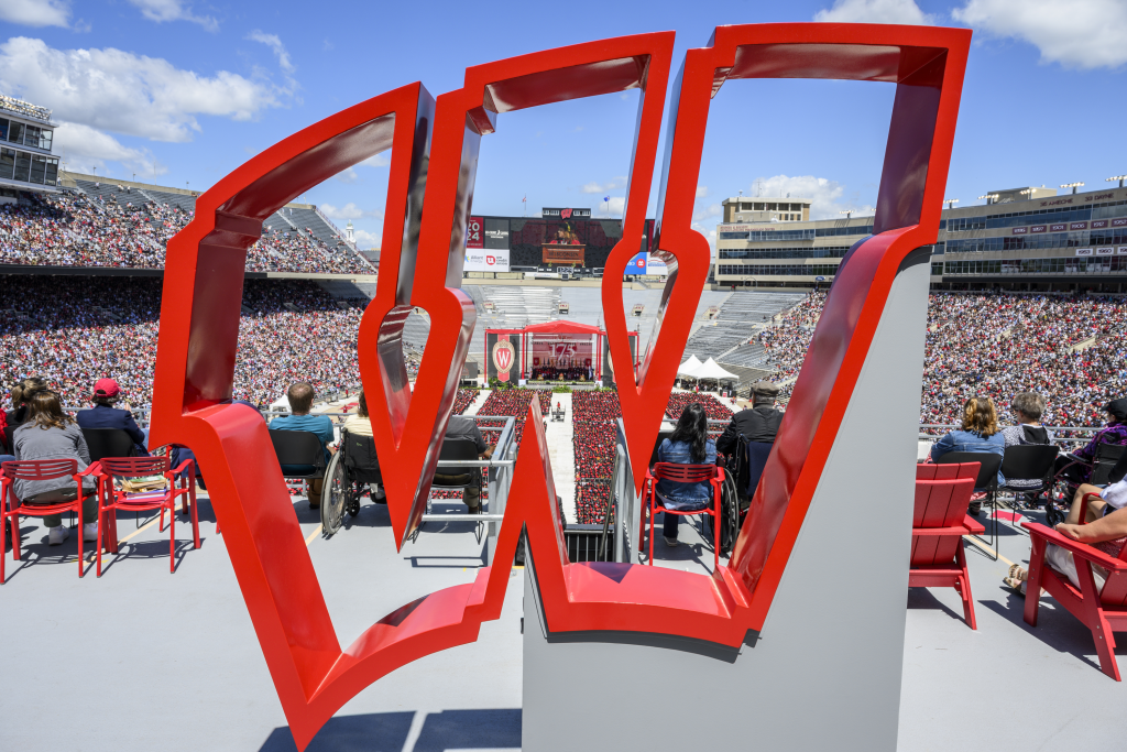 Photo of hollow metal sculpture of action W letter overlooking Camp Randall stadium full of UW graduation attendees and graduates.