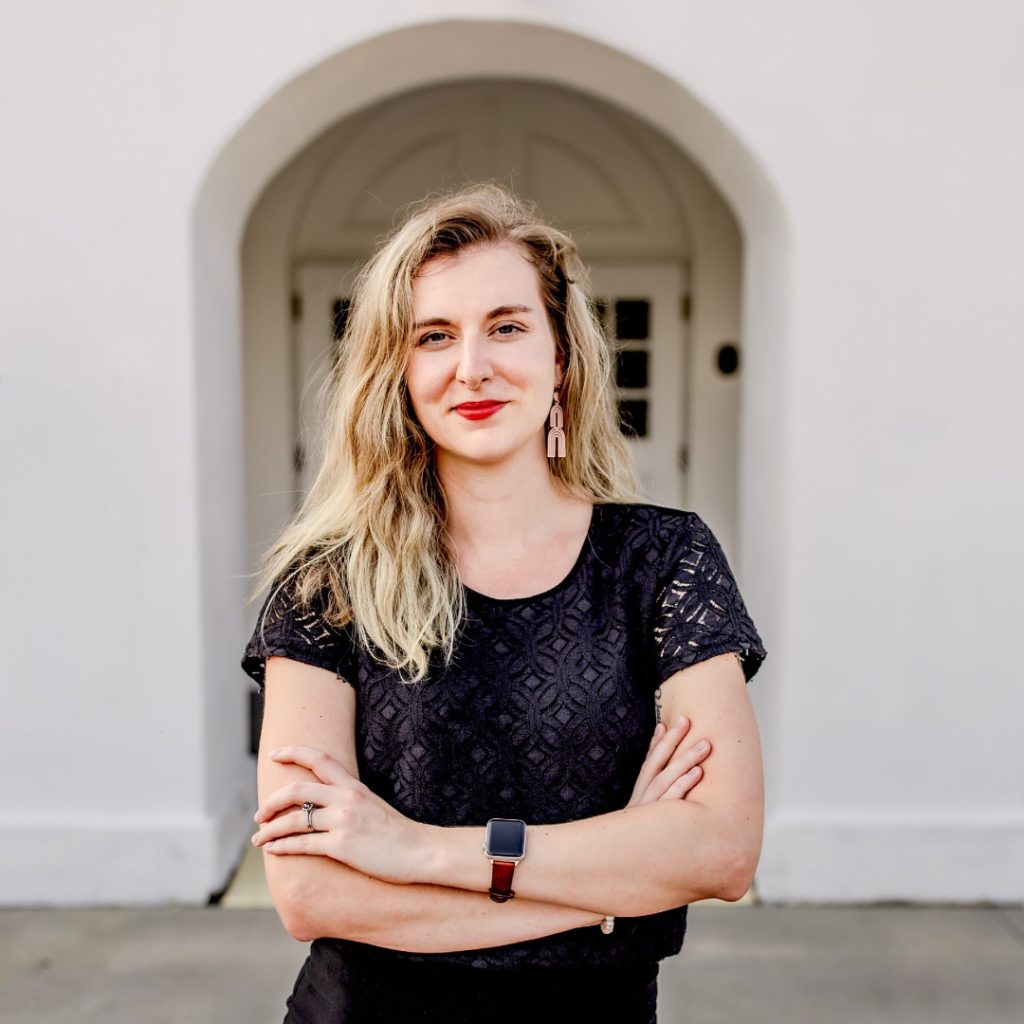 Portrait of Alisha Esselstein '18, a young woman with fair skin, blonde hair that is a bit more than shoulder length, red lipstick, and is wearing a black patterned top while standing with arms crossed in a confident expression.