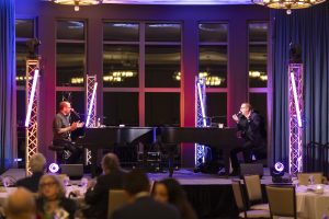 Dueling pianists converse on stage at their pianos while a crowd watches on and pink and purple lights illuminate everything.