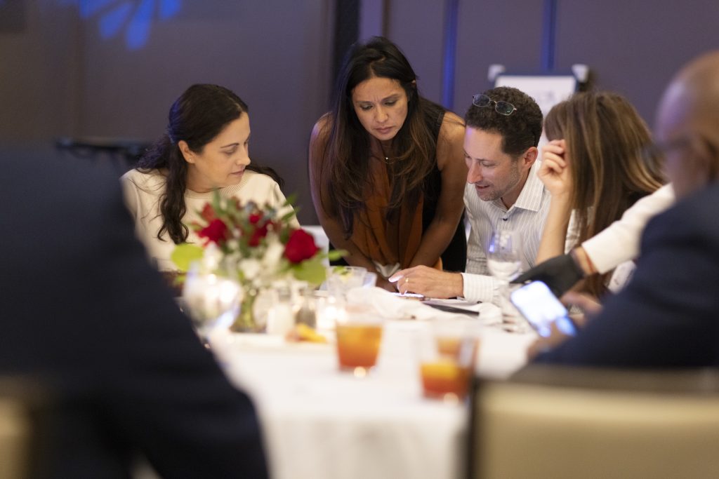 Photo of people socializing at a reunion. A few people look together at a man's phone on a table.