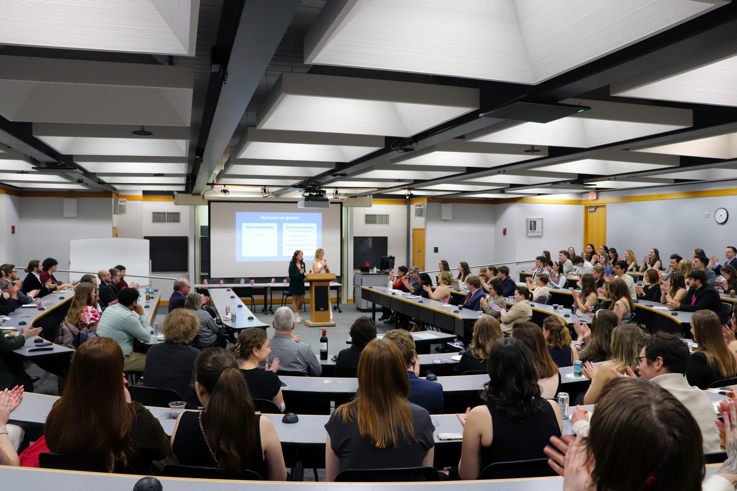 A photo of the Jackie Macaulay Auction, showing a room packed with attendees in a semicircle with rows of tables, and two women presenters at the center.