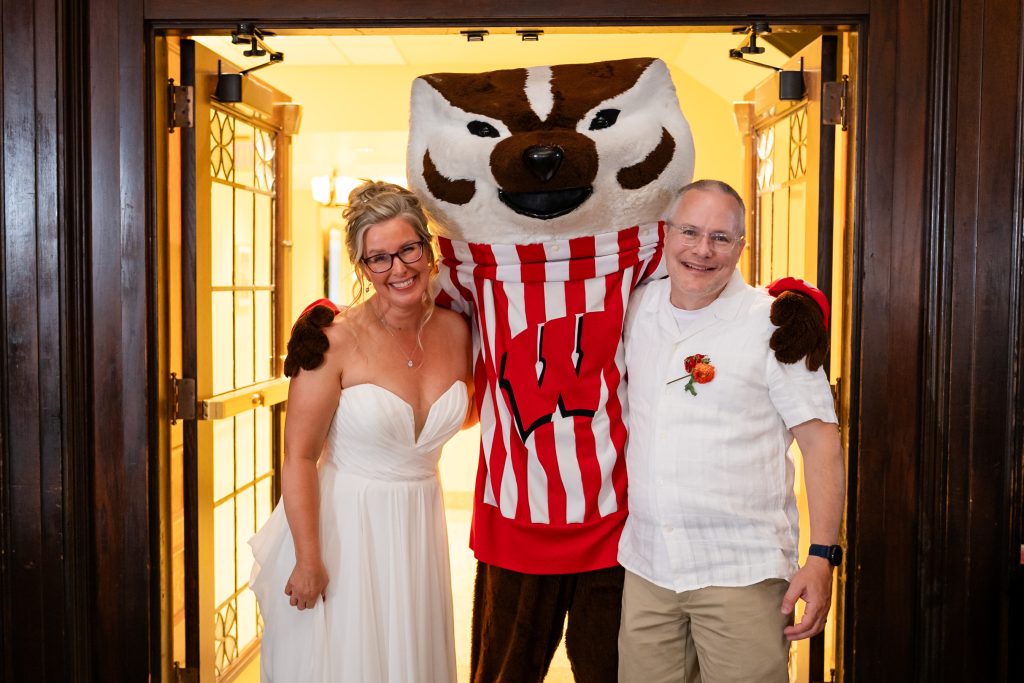Photo of Sara and Dave Waters on their wedding day while being hugged by someone in a large Bucky Badger costume.