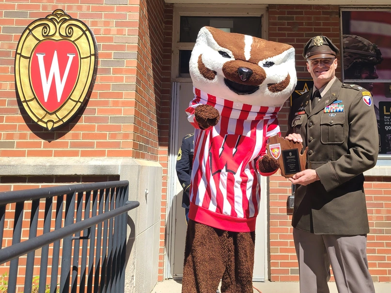 A man in military uniform accepts a wooden plaque from someone Dressed in full Bucky Badger costume.