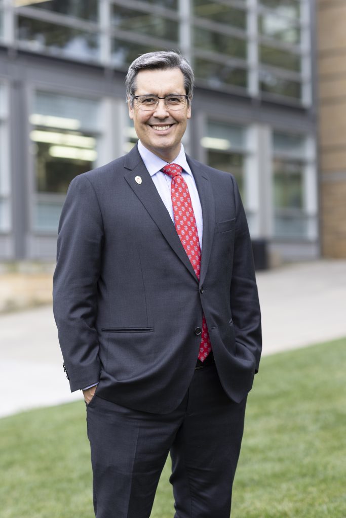 Dean Dan Tokaji stands with a smile while wearing a grey suit with red and white gargoyle styled tie. He stands in front of the UW Law School building.