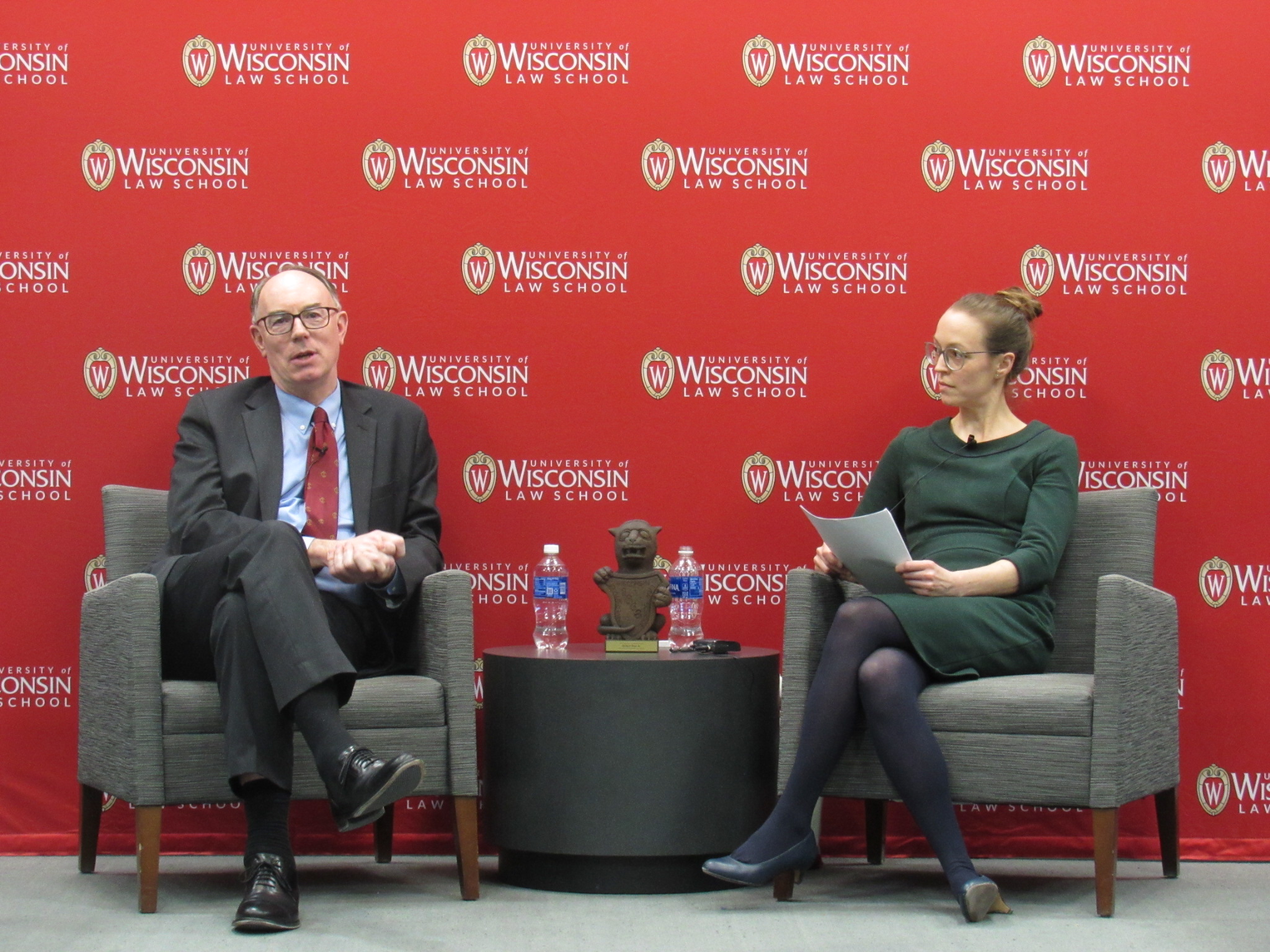 The Fairchild Lecture speaker, Honorable Robert M. Dow, Jr. speaks with Nina Varsava on stage. They're both seated in fabric chairs with a Gargoyle statue in between.
