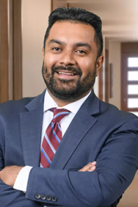 Photo of Tomás Clasen, a man of color with a light beard, blue suit, white shirt, and red striped tie, while smiling at the camera.