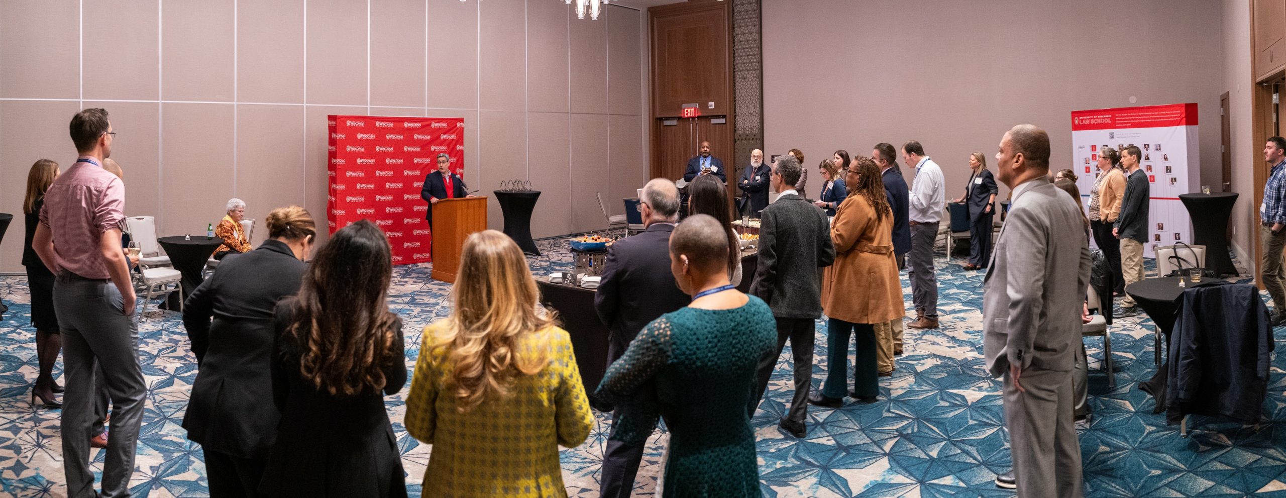 Numerous people stand in the event space to listen to Dan Tokaji speaking.