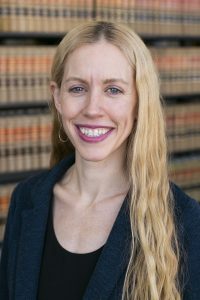 Photo of a fair skinned woman with long, wavy blonde hair and blue eyes. She is wearing a blue/gray blazer over a black top.