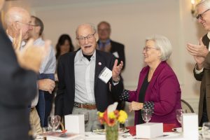an elderly man smiles as many people applaud to him