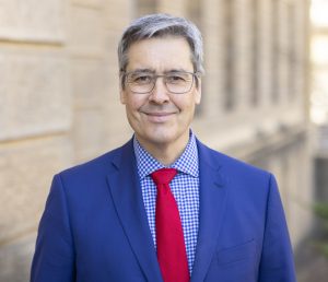 Photo of Dan Tokaji, a gray haired man of asian descent wearing glasses, a bright blue suit and red tie, and a checkered blue shirt beneath the jacket.