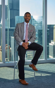 A young black man with a trim beard wears business attire and sits on a stool that is in front of a window with a city skyline outside.